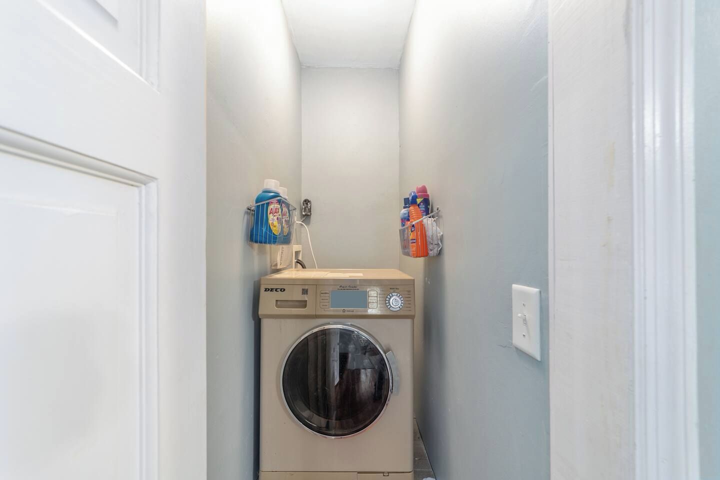 Une buanderie équipée d’un lave-linge DECO, d’un distributeur de lessive et d’un étagère contenant des produits de nettoyage.
