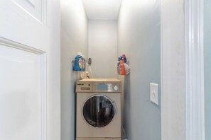 A laundry room with a DECO washing machine, a detergent dispenser, and a rack with cleaning supplies. - Martin House | Historic Suite with Pool Downtown (Ocala)