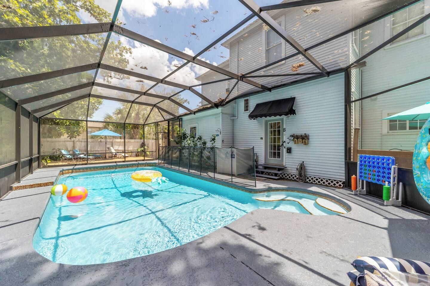 A covered outdoor swimming pool with a glass enclosure, a lounge area with chairs, and a house in the background.