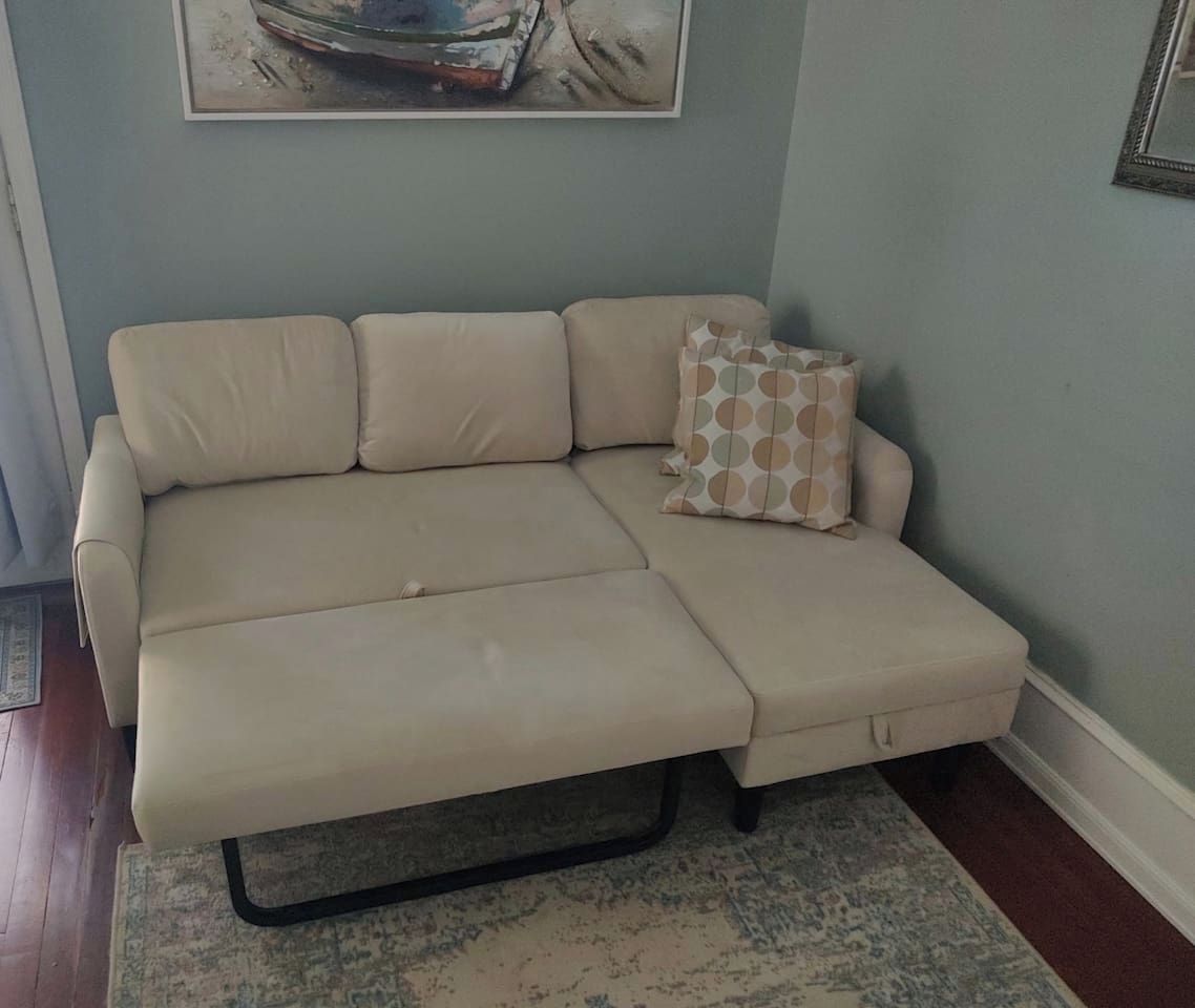 A beige sectional sofa with a matching ottoman, a patterned throw pillow, and a framed artwork above it.