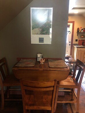 A dining area with a wooden table and chairs, a framed picture of a landscape, and a kitchen visible through an open doorway.
