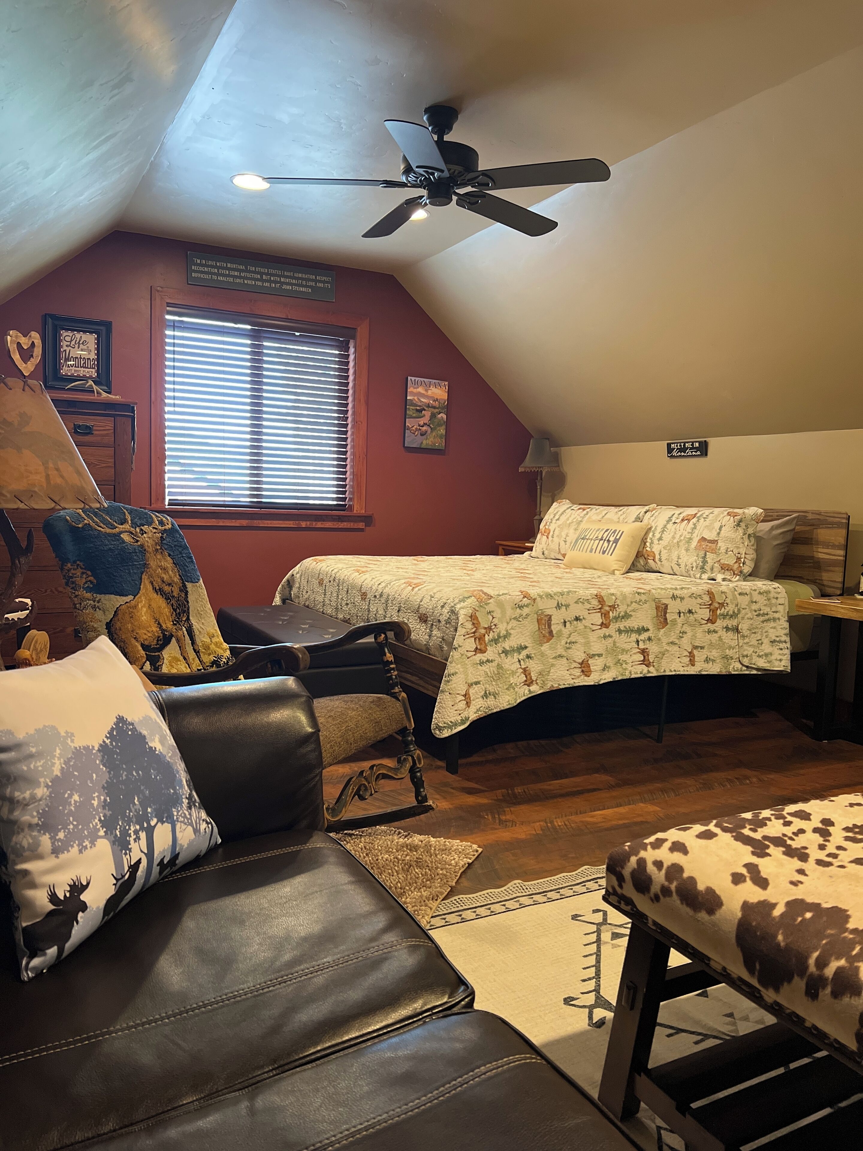 Ein gemütliches Schlafzimmer im Dachgeschoss mit schrägem Dach, Deckenventilator, Bett mit Blumenmusterdecke, Ledersofa und Sessel mit Kissen mit Hirschmotiv.