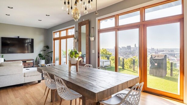 Ein geräumiger Essbereich mit Holztisch und weißen Stühlen, großen Fenstern mit Blick auf die Stadt und einem an der Wand befestigten Flachbildfernseher.