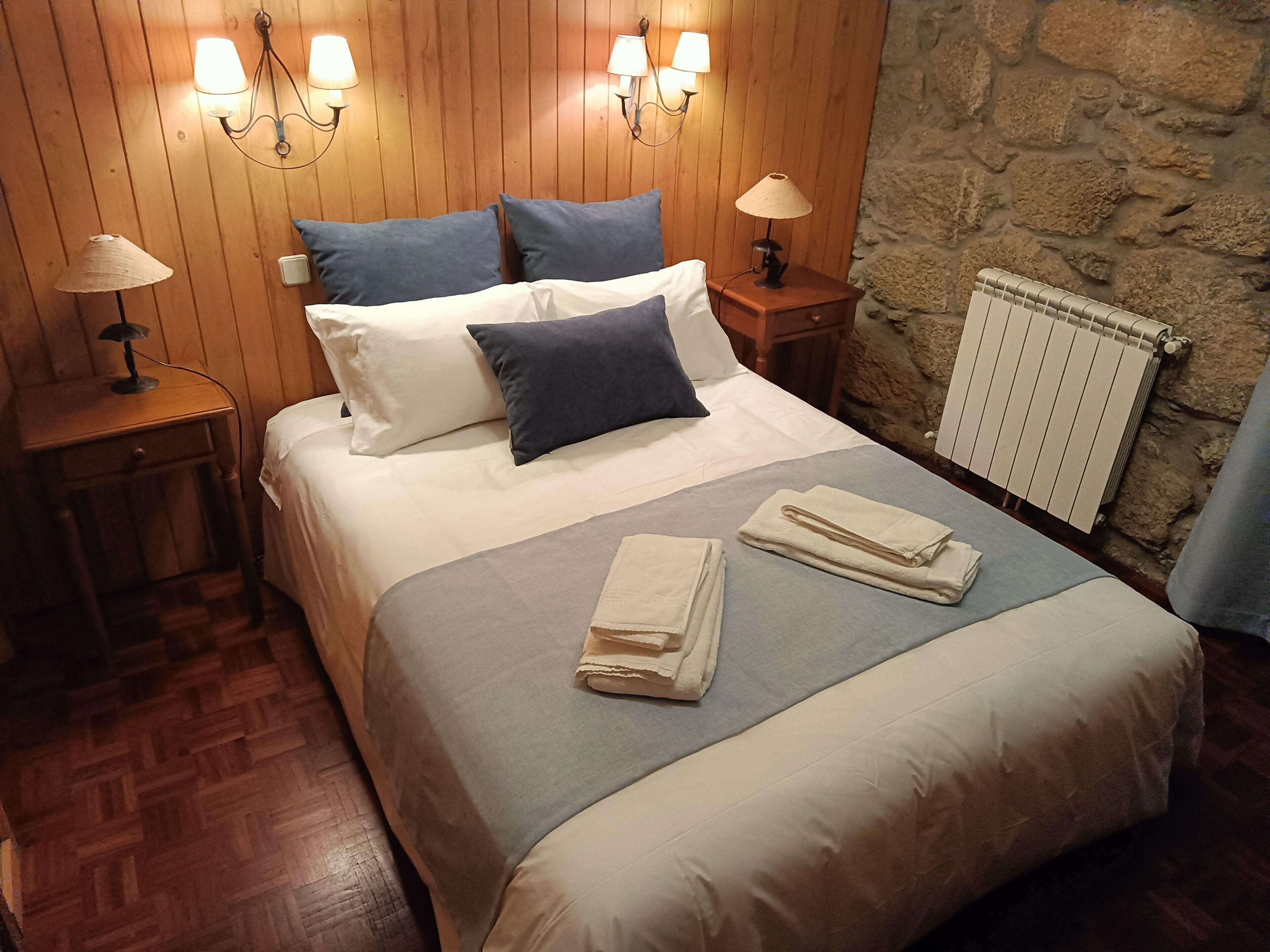 A neatly made bed with pillows, a bedside table with a lamp, and a stone wall.
