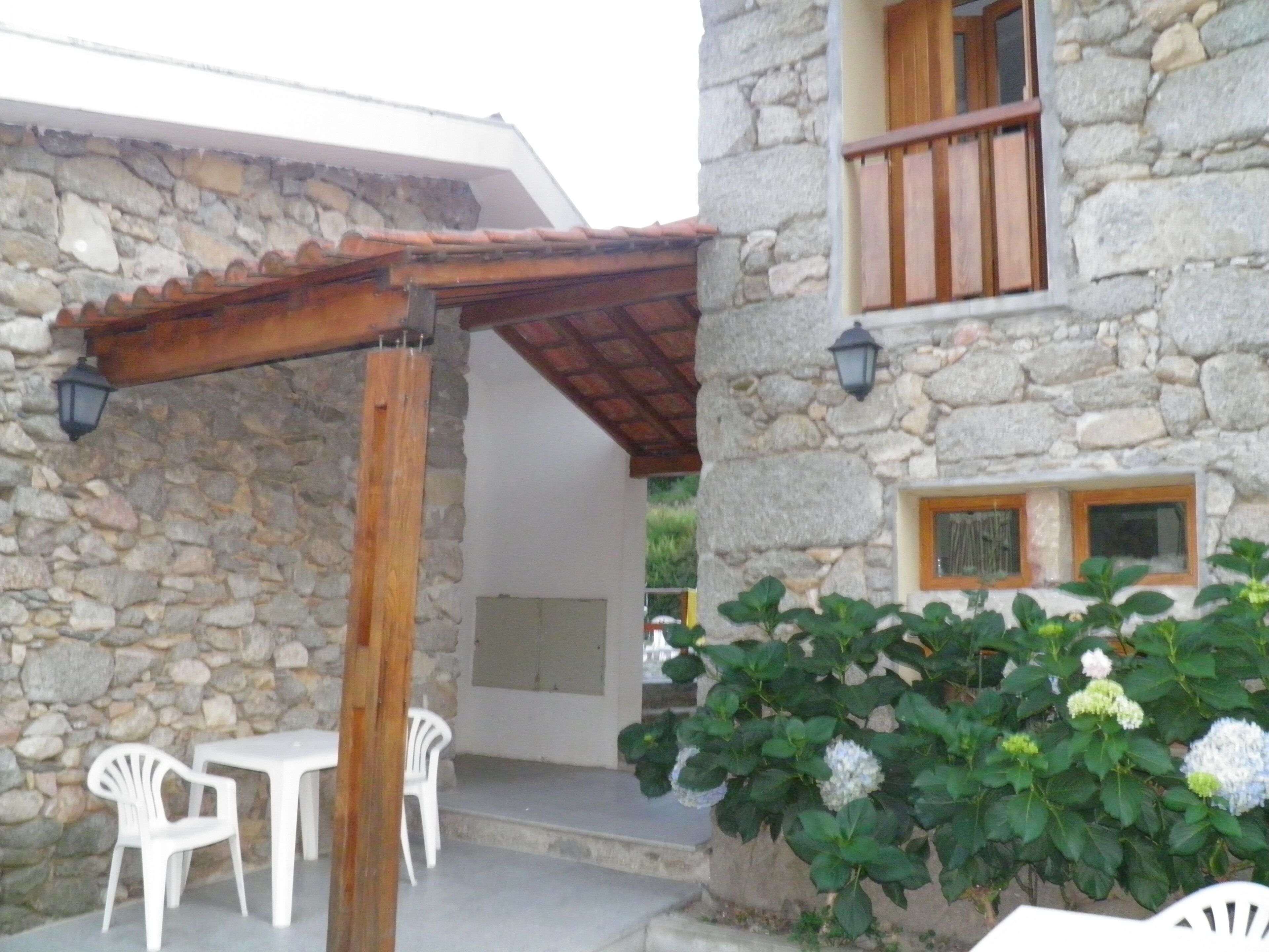 A stone building with a wooden pergola, white plastic chairs, and a plant with white flowers.
