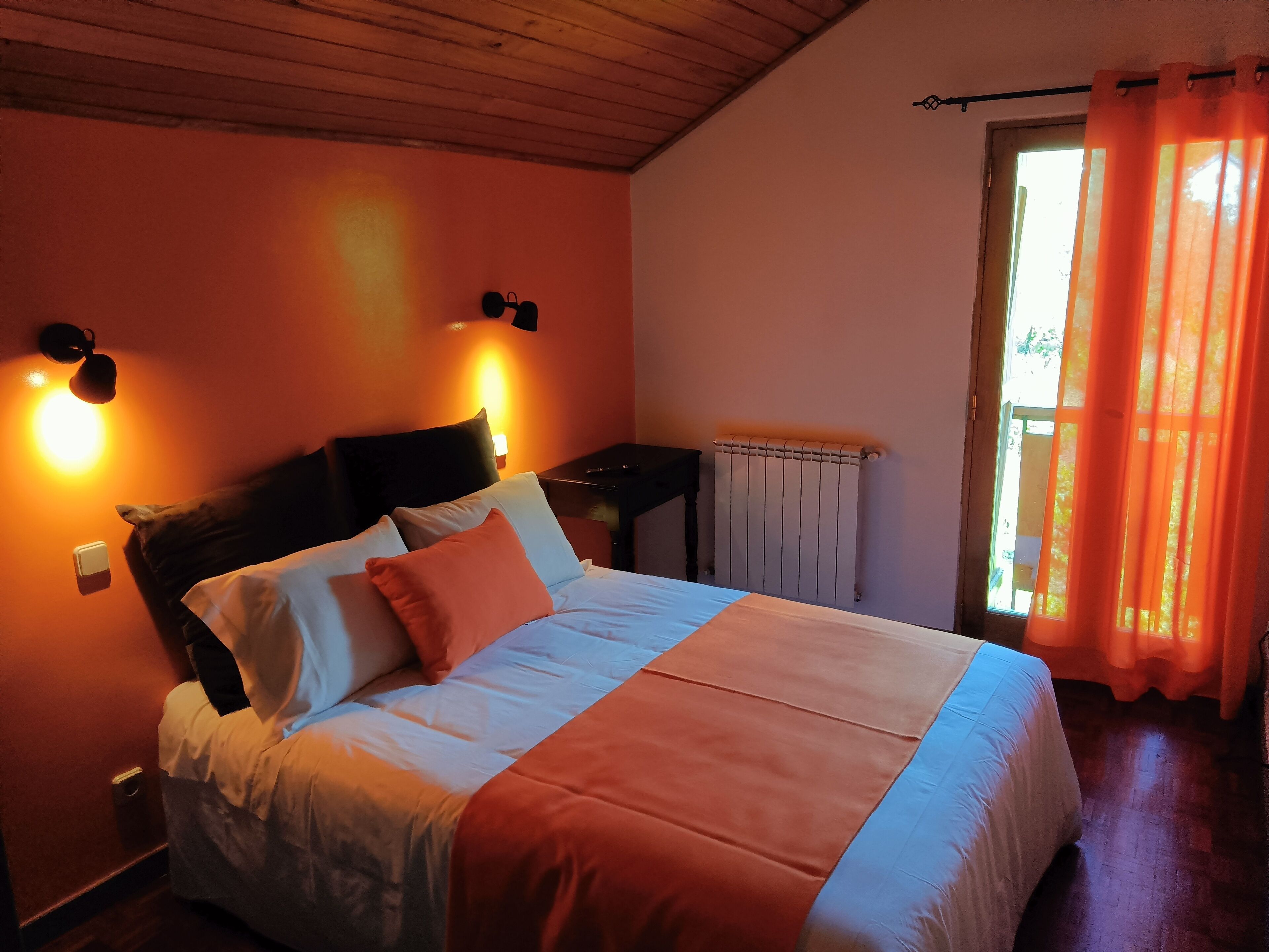 A bedroom with a bed, a nightstand, a wall-mounted lamp, a radiator, and a window with curtains.