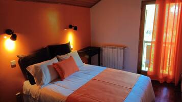 A bedroom with a bed, a nightstand, a wall-mounted lamp, a radiator, and a window with curtains.