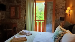A room with a bed, a wooden headboard, a nightstand, a lamp, a window with curtains, and a view of greenery outside. - Quinta de Leandres (Manteigas)