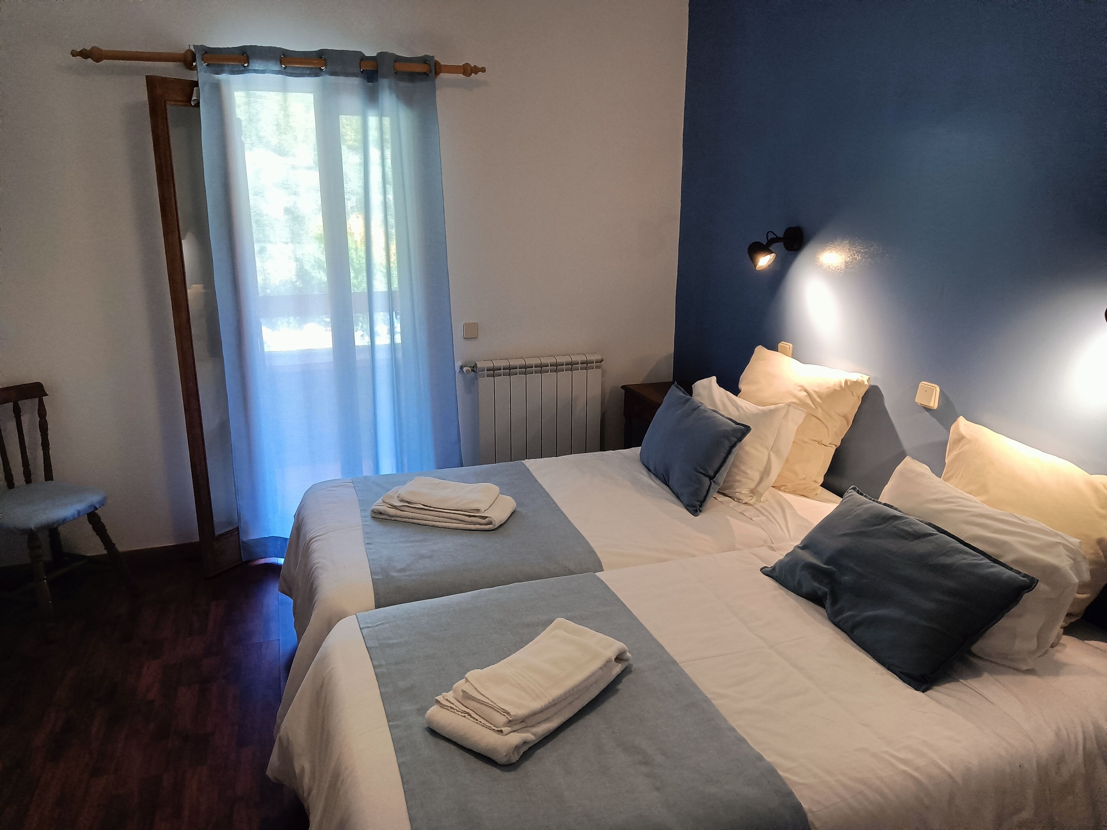 A hotel room with two beds, a chair, a radiator, and a window with curtains.