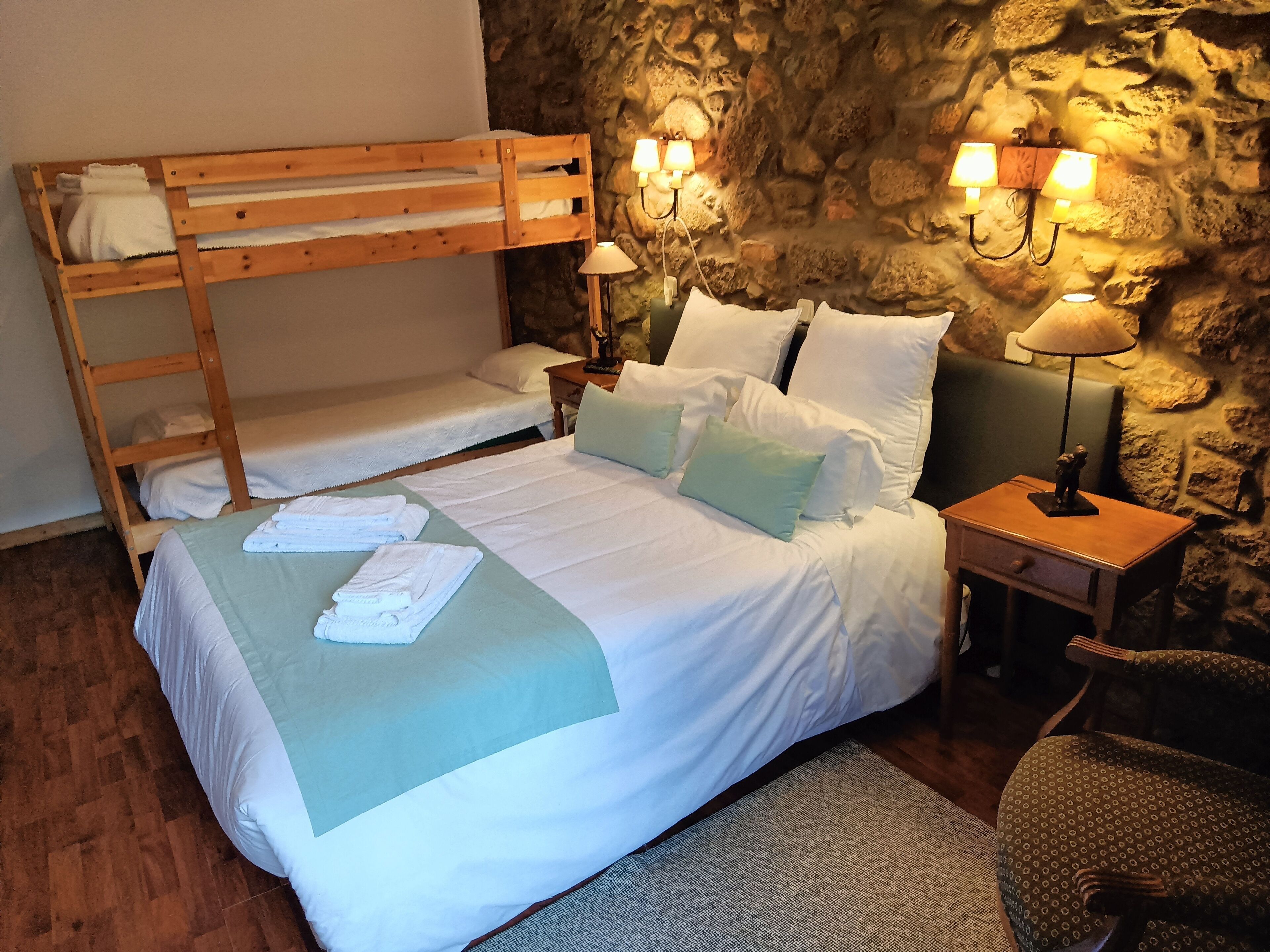 A bunk bed room with a stone wall, two bedside lamps, a small table with a statue, and a chair.
