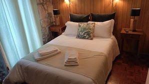 A neatly made bed with white linens and a tropical leaf pillow, flanked by two nightstands with lamps, in a room with wooden paneling and a stone accent wall. - Quinta de Leandres (Manteigas)