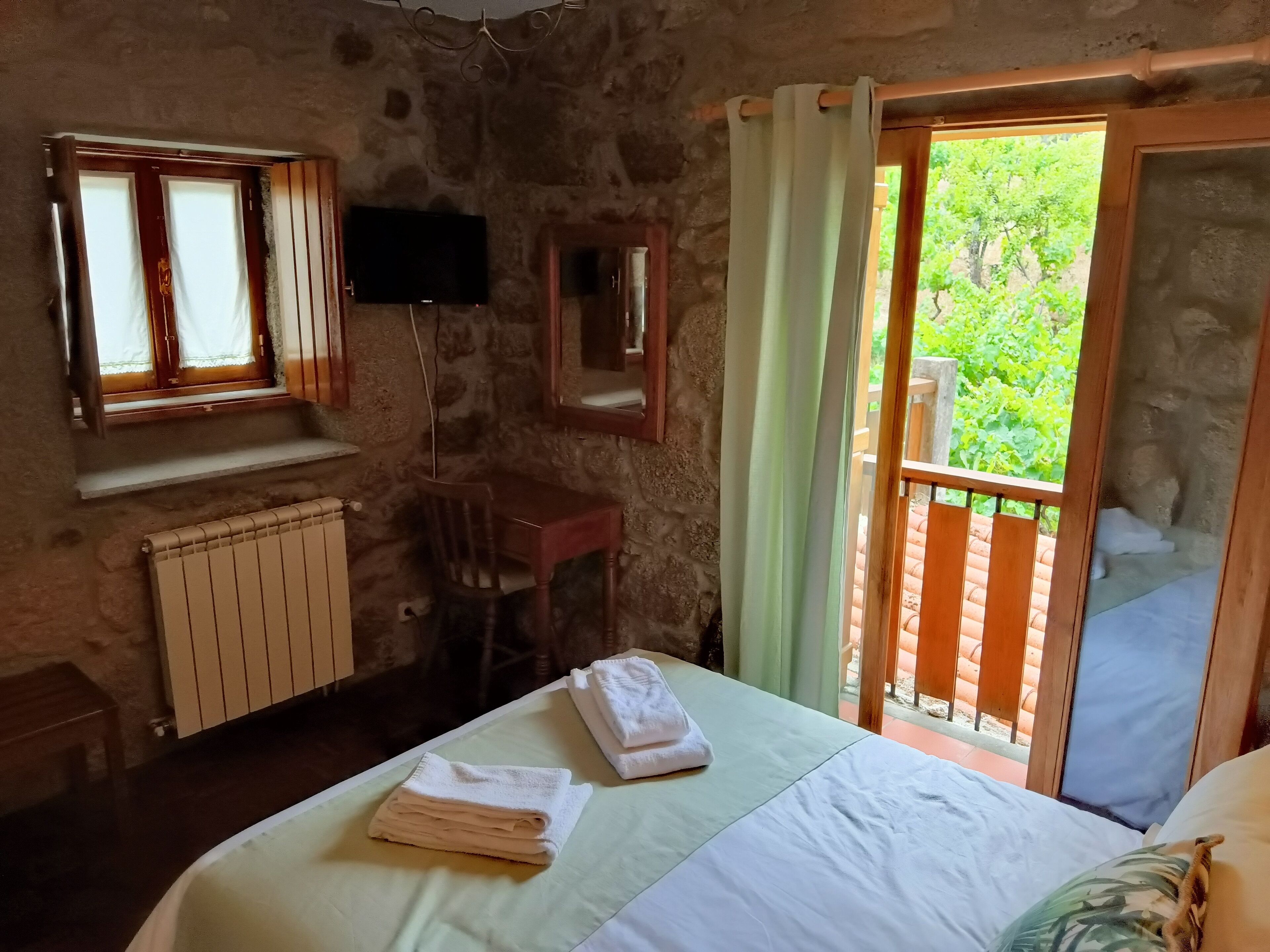 A room with a bed, a chair, a desk, a radiator, a TV, and a balcony with a view of greenery.