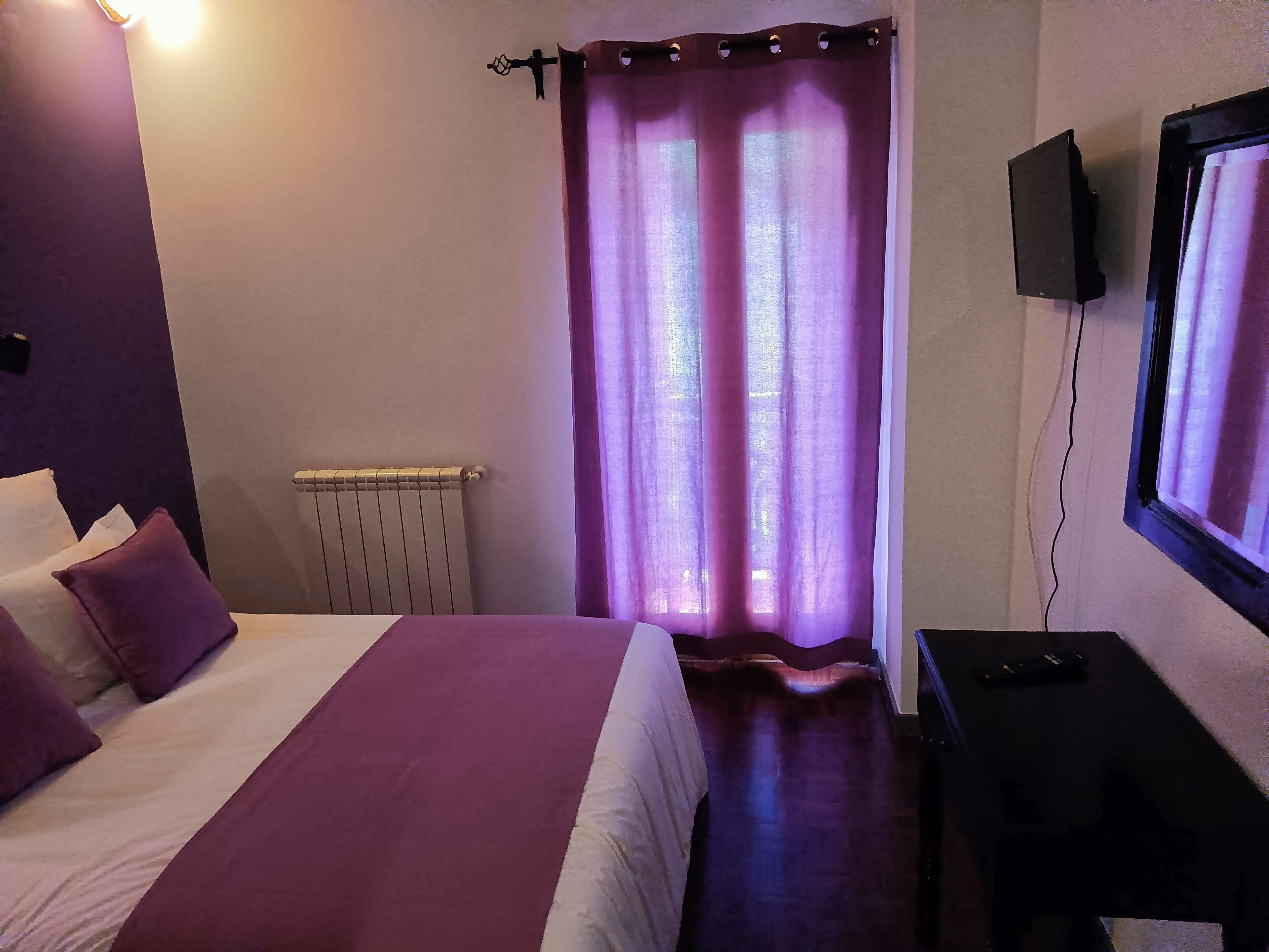 A hotel room with a bed, a radiator, a TV, and purple curtains.
