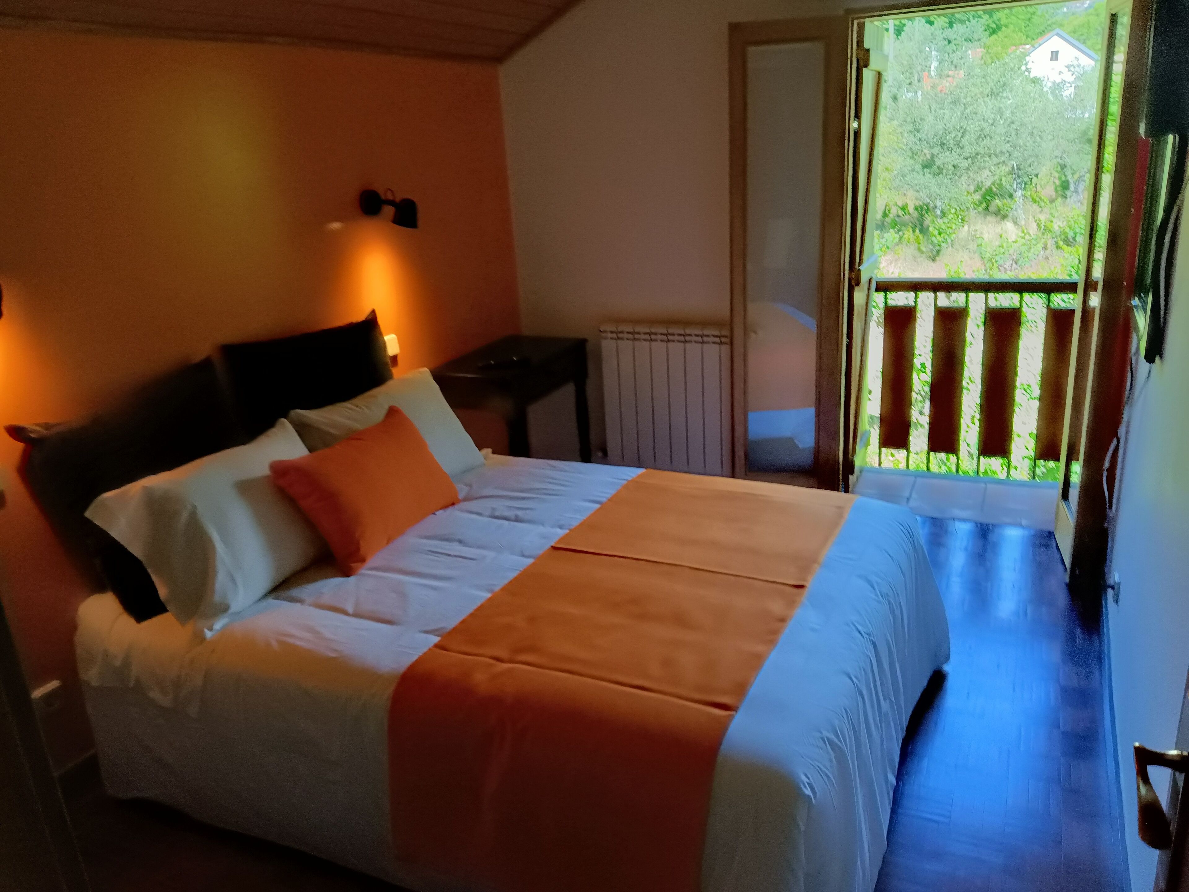 Un dormitorio con cama, almohadas, una mesita de noche, una ventana que da a zonas verdes y una puerta que sale al exterior.