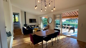 A modern living room with a wooden floor, a sofa, a dining table with chairs, a TV mounted on the wall, and a sliding glass door leading to an outdoor patio.