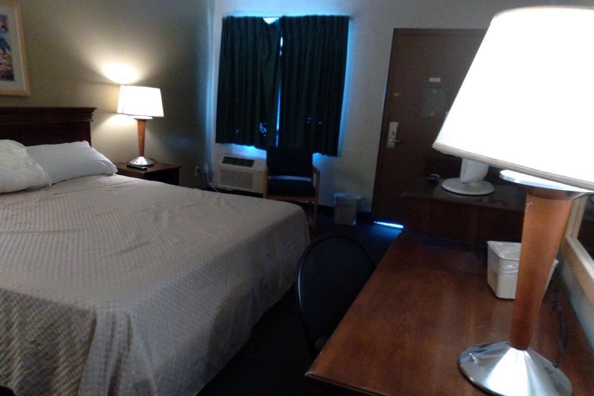 A hotel room with a bed, a desk, a chair, a lamp, a window with curtains, and a door.