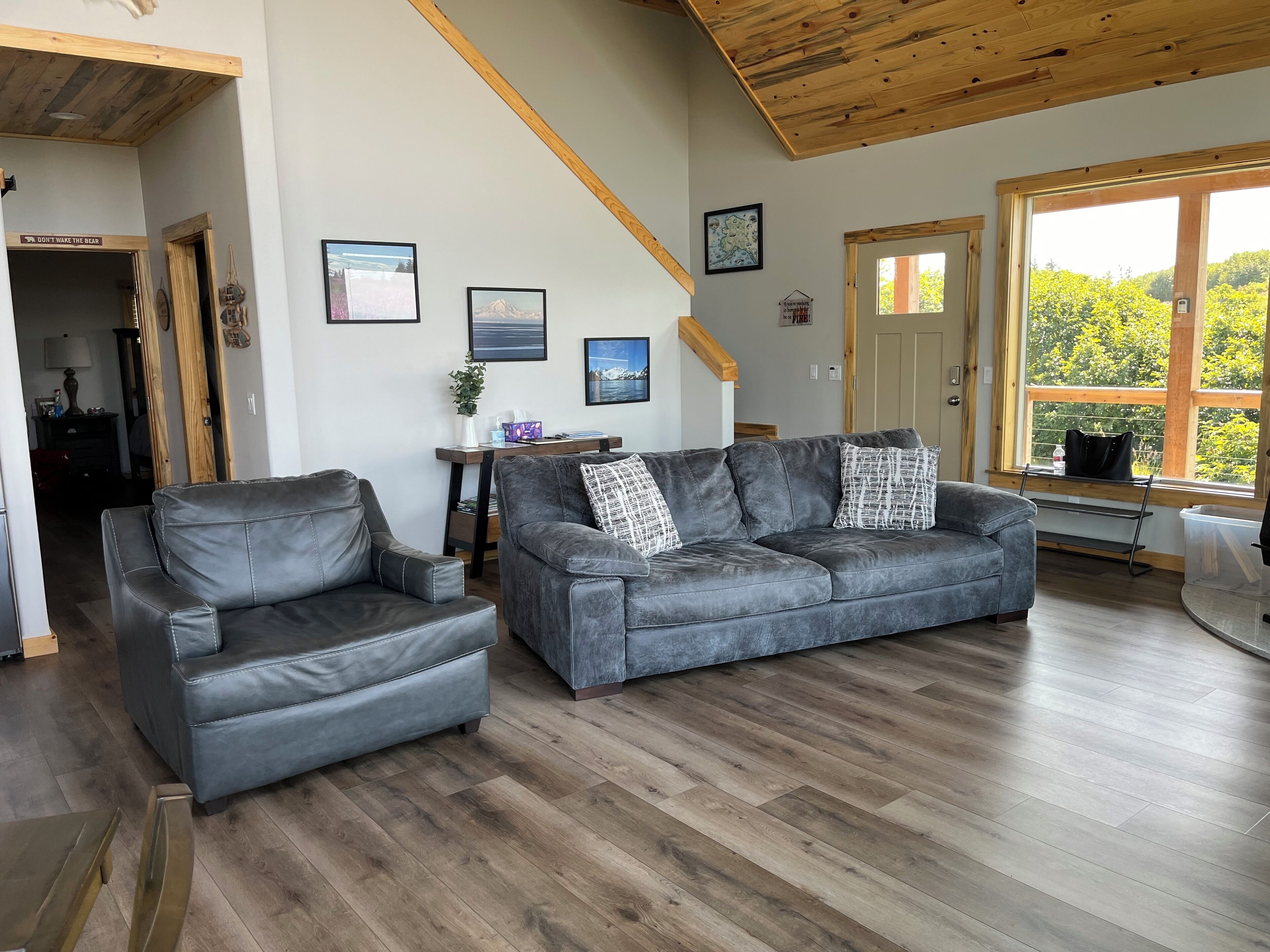 A modern living room with a grey leather sofa, a matching armchair, and a wooden ceiling. There are framed pictures on the wall, a small table with a plant, and a window with a view of greenery.