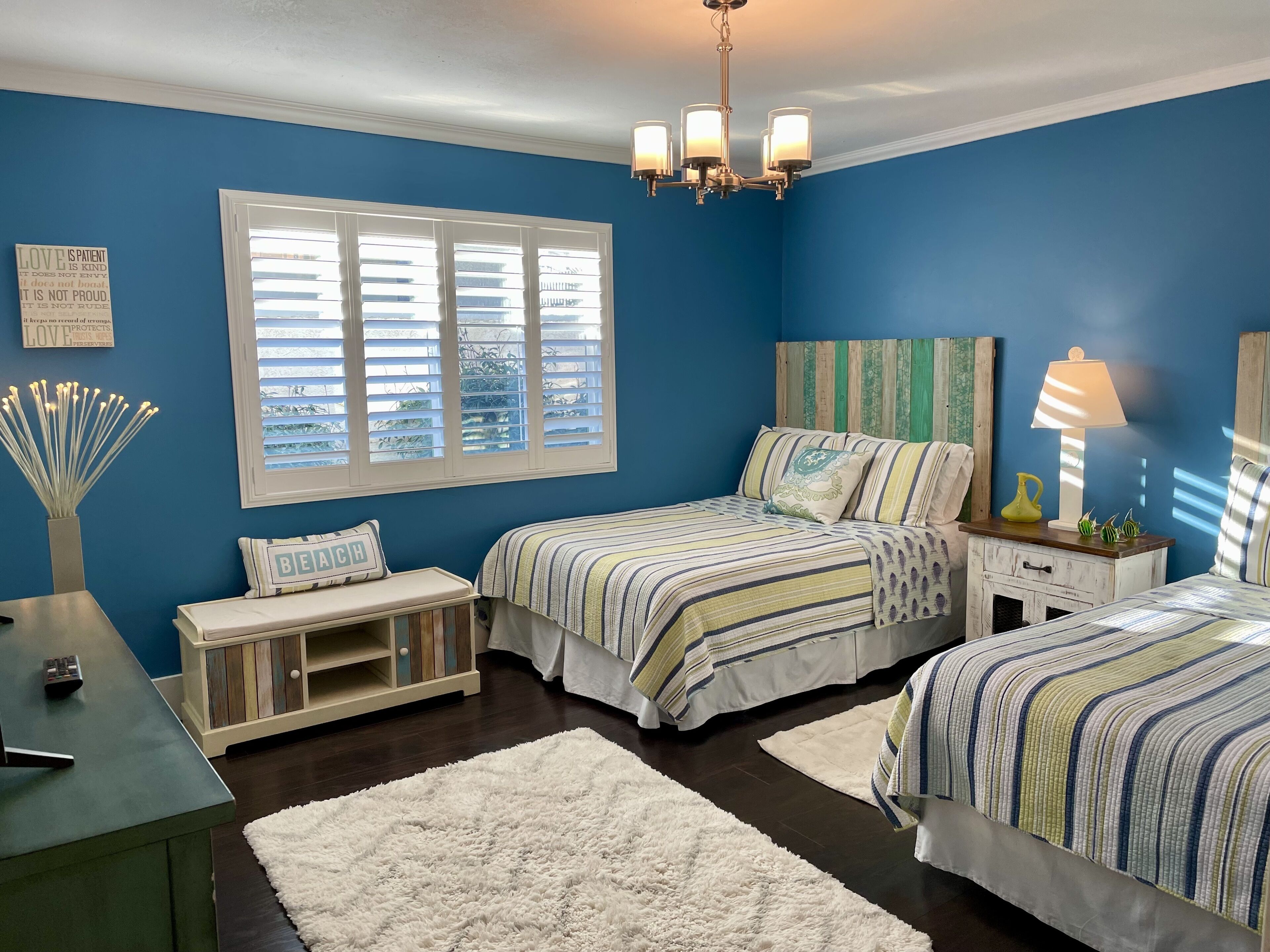 Quarto com duas camas, janela com persianas, lustre e mesa de cabeceira com abajur.