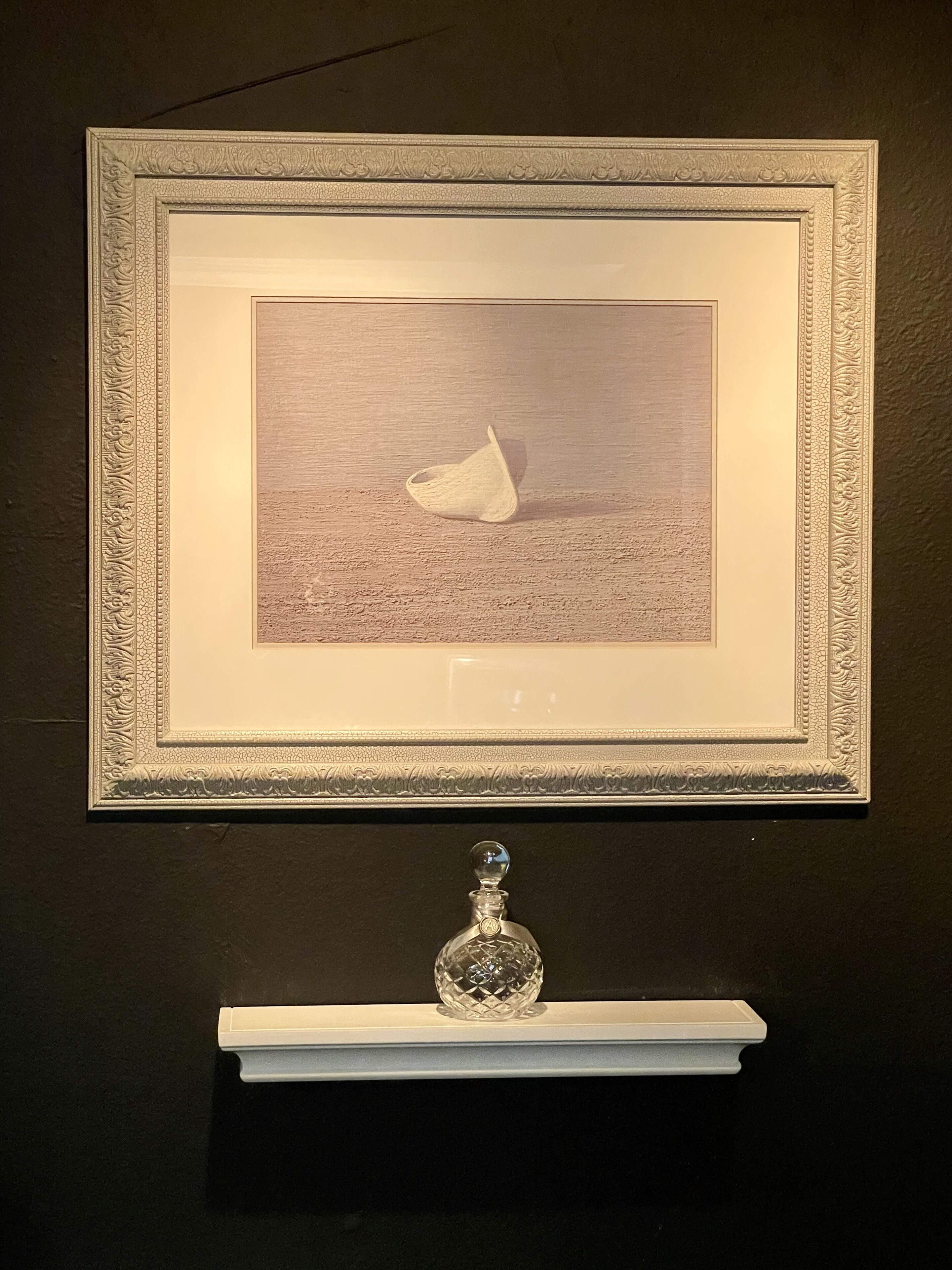 A framed artwork of a boat on water, displayed on a wall with a decorative border. Below the artwork, there is a white shelf with a crystal bottle.