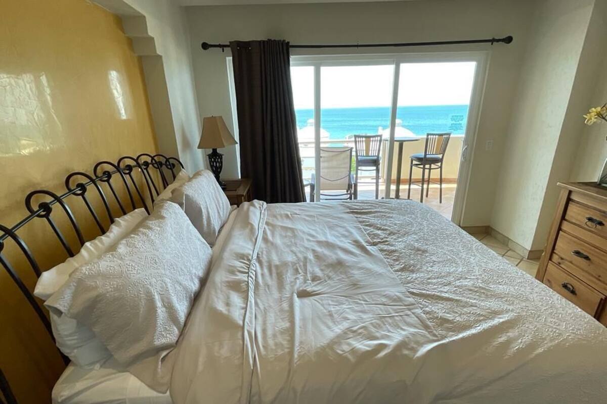 A hotel room with a bed, a nightstand, a lamp, a dresser, and a view of the beach and ocean through a large window.