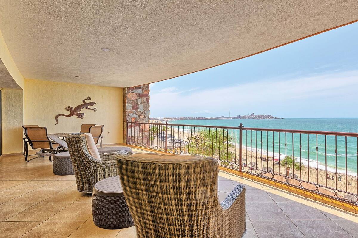 A balcony with wicker furniture, a stone wall, and a view of the sea and distant cityscape.