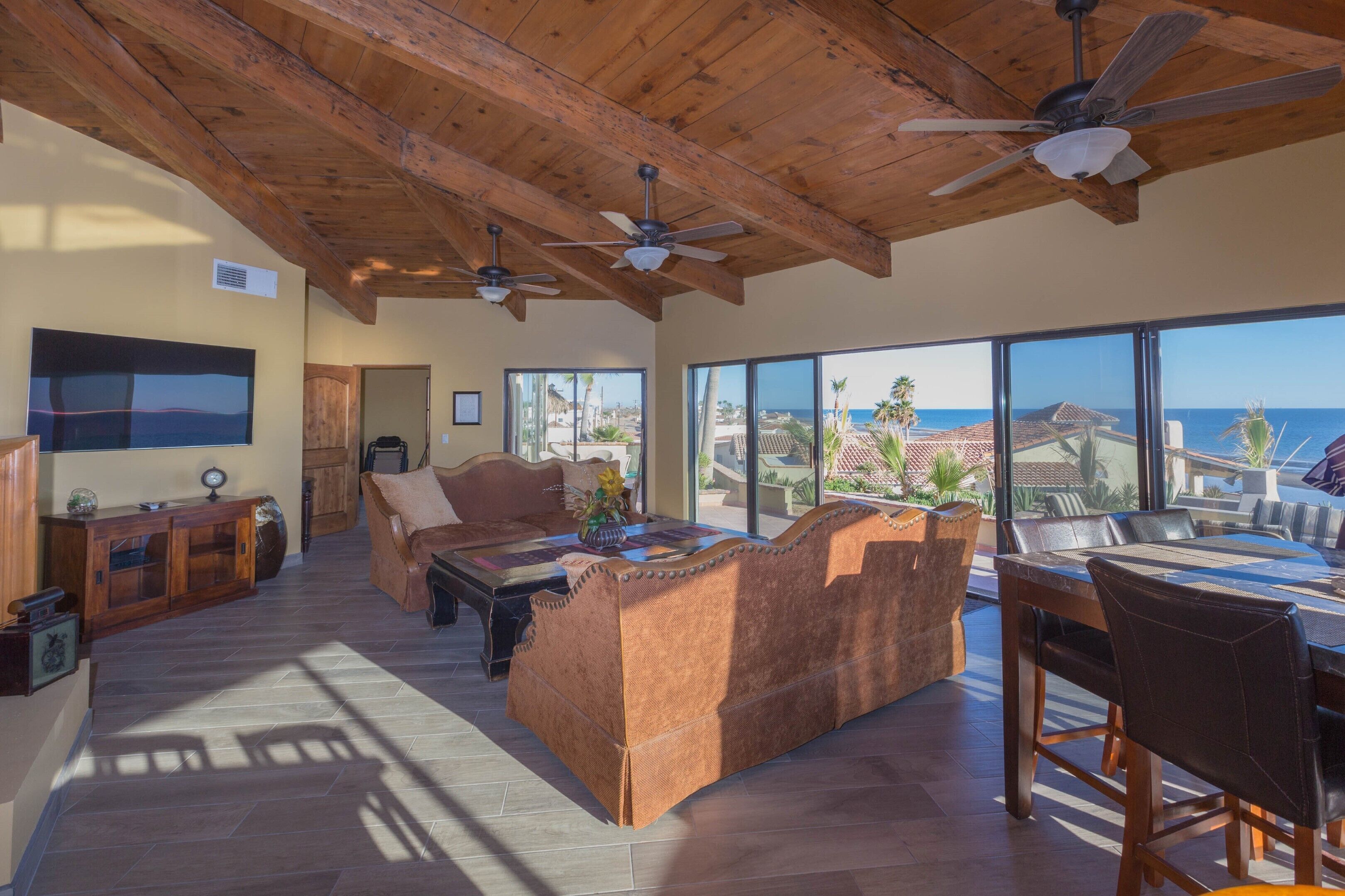 A spacious living area with a wooden ceiling, large windows offering a view of the ocean, a sofa, and a dining table.