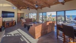 A spacious living area with a wooden ceiling, large windows offering a view of the ocean, a sofa, and a dining table. - RPB Property Management (Puerto Peñasco)