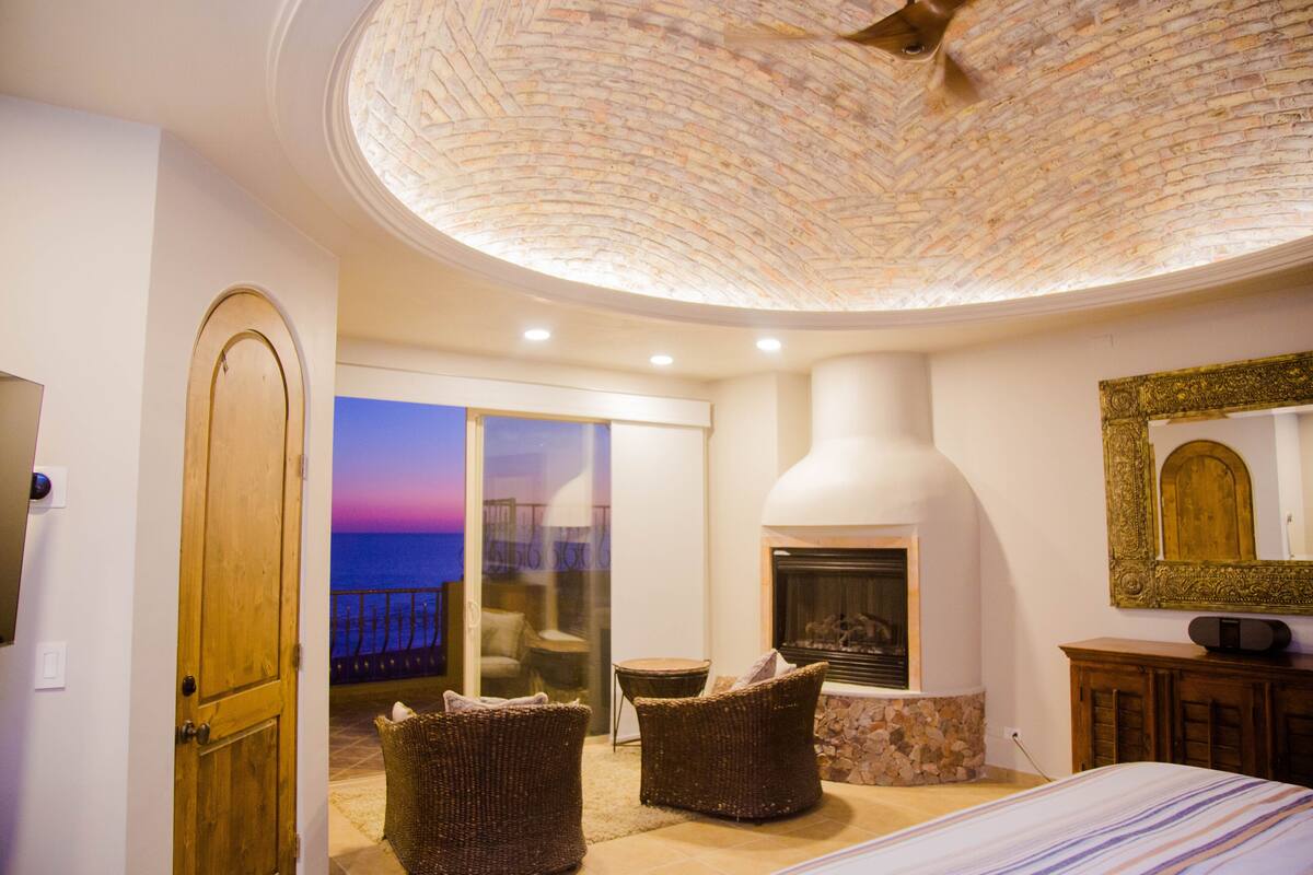 A room with a fireplace, wicker chairs, a bed, and a view of the sea.