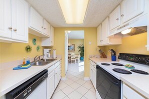 Cocina con alacenas blancas, estufa y lavavajillas. Se ve un comedor a través de un vano abierto.