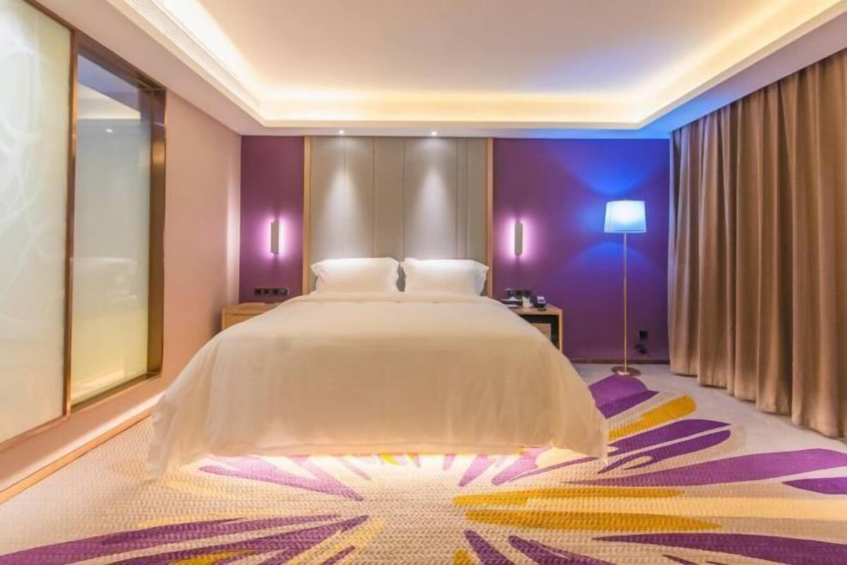 A hotel room with a large bed, a nightstand, a lamp, and a view of the outside.