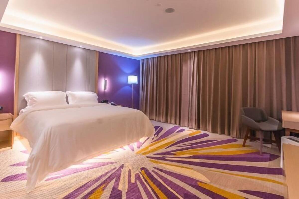 A hotel room with a bed, a desk, a chair, a large window with curtains, and a patterned rug.