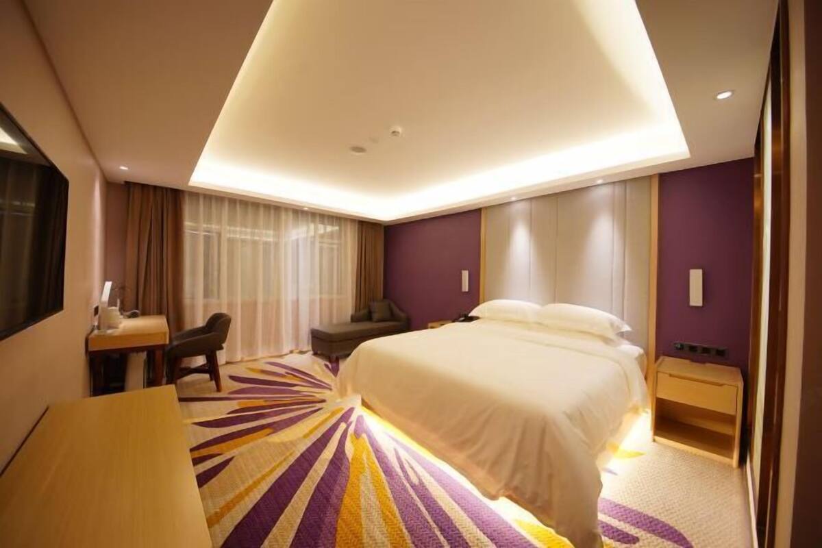 A hotel room with a large bed, a desk with a chair, a small table, and a bedside table.