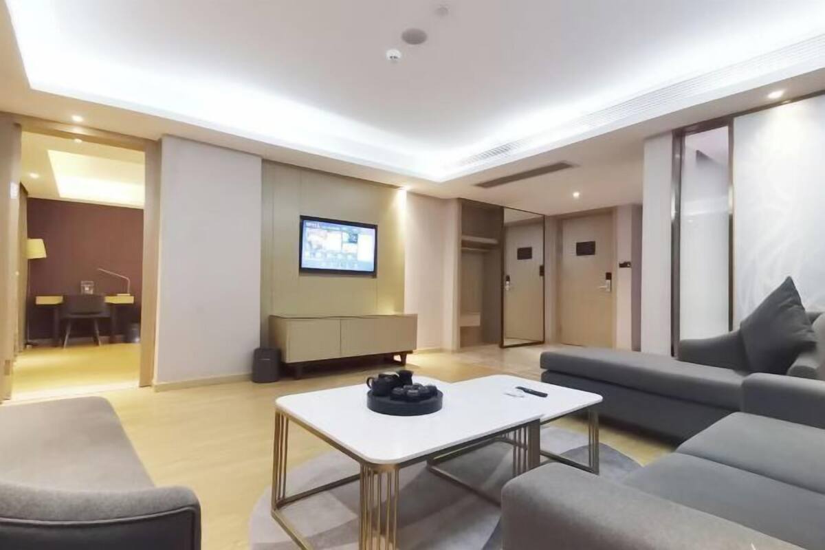 A modern hotel room with a sofa, a small table, a TV mounted on the wall, and a door leading to another room.