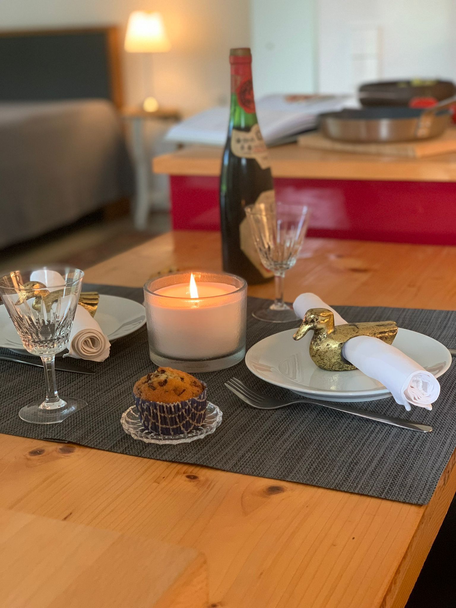 A dining table with a candle, a glass of wine, a gold swan ornament, a rolled napkin, a muffin, and a glass of water.
