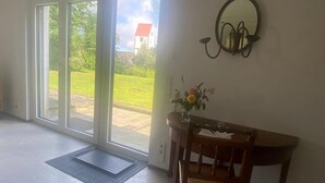 Huoneessa on liukuovet, jotka johtavat puutarhaan, puinen tuoli, pieni pöytä, jossa on maljakko täynnä kukkia, ja seinälle kiinnitetty peili.