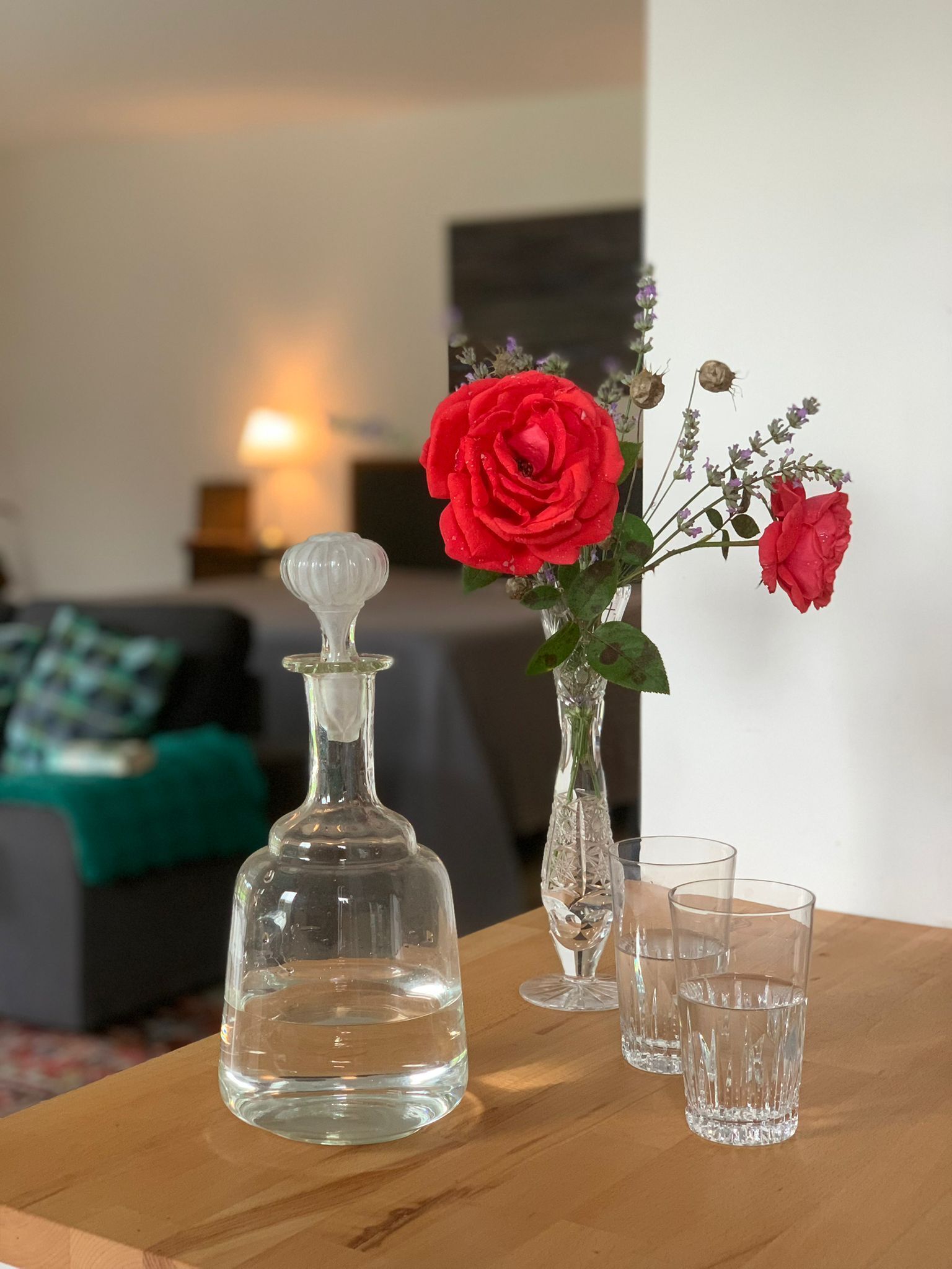 A clear glass decanter, a vase with red roses, and three clear glasses on a wooden table.