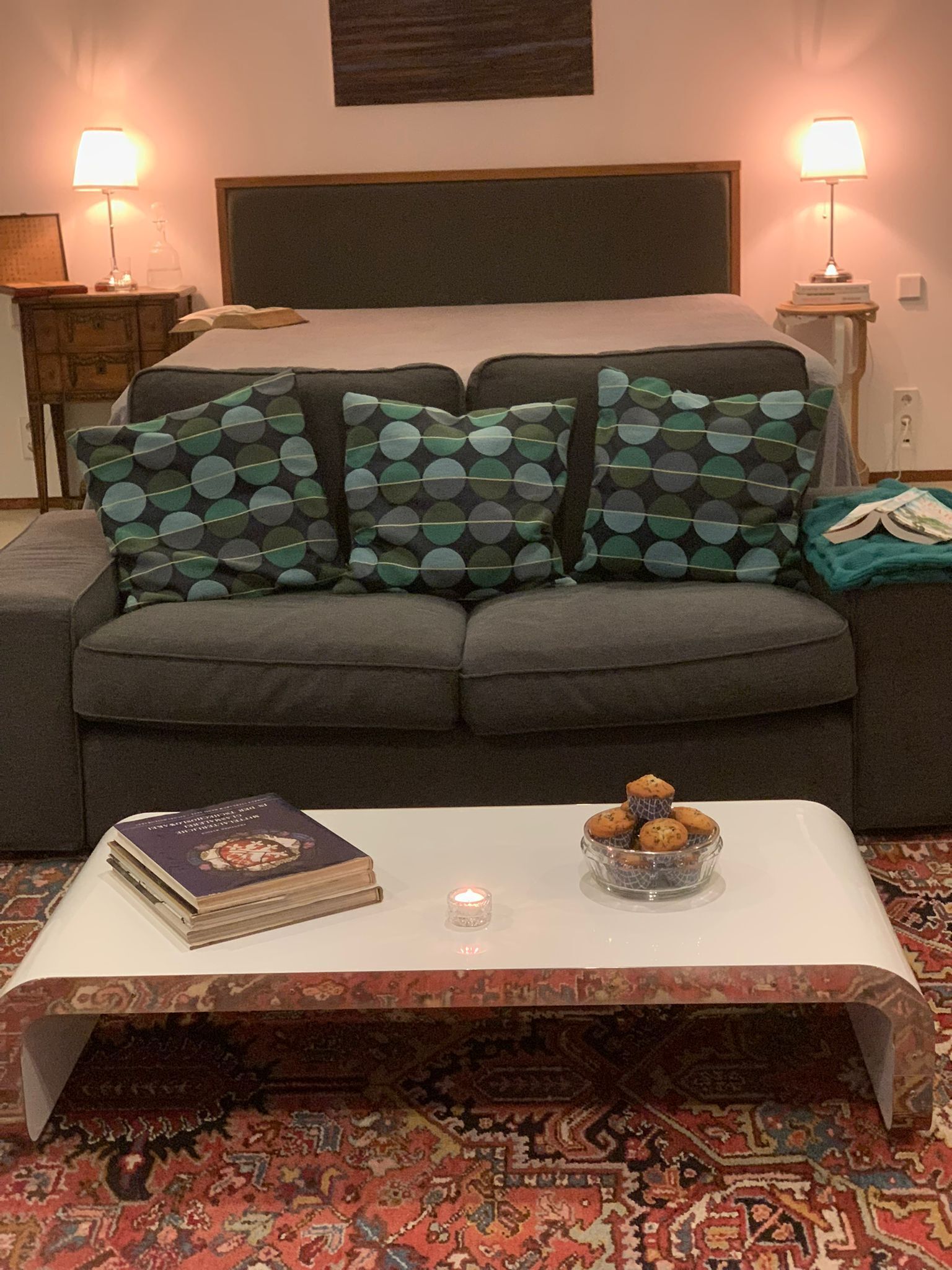 Ein gemütliches Wohnzimmer mit grauem Sofa, gemusterten Kissen, einem Couchtisch mit Buch und Kerze sowie einem Teppich mit Muster.