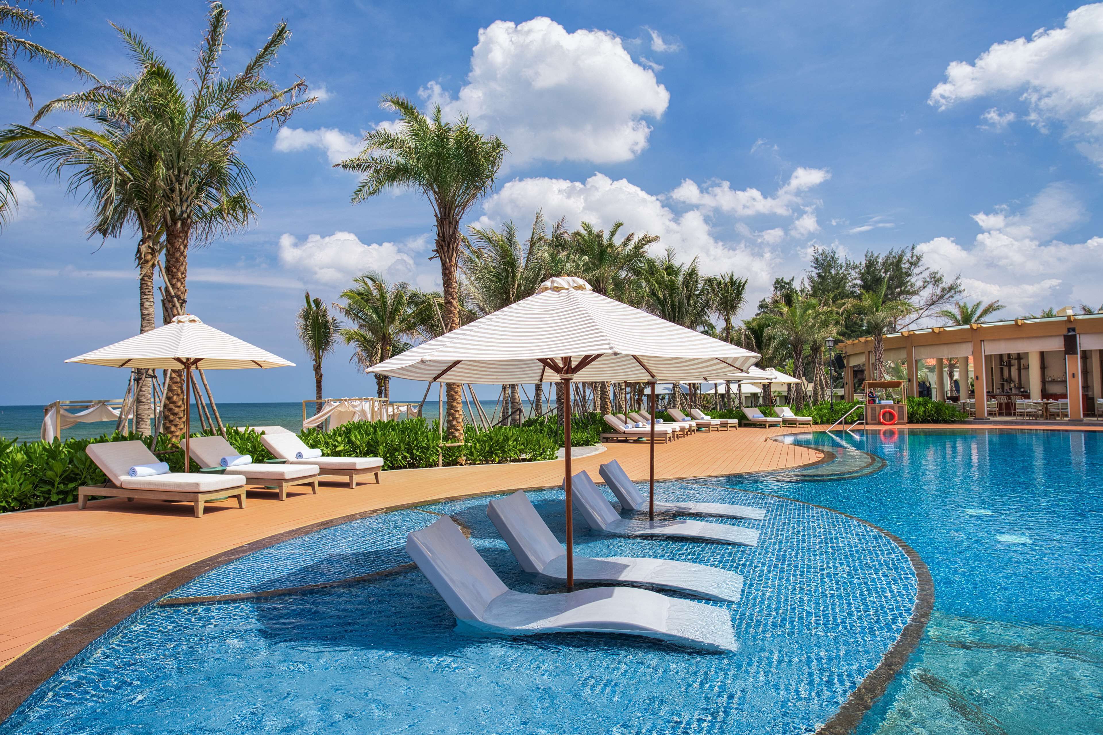 A pool area with lounge chairs and umbrellas, surrounded by palm trees and a clear sky.
