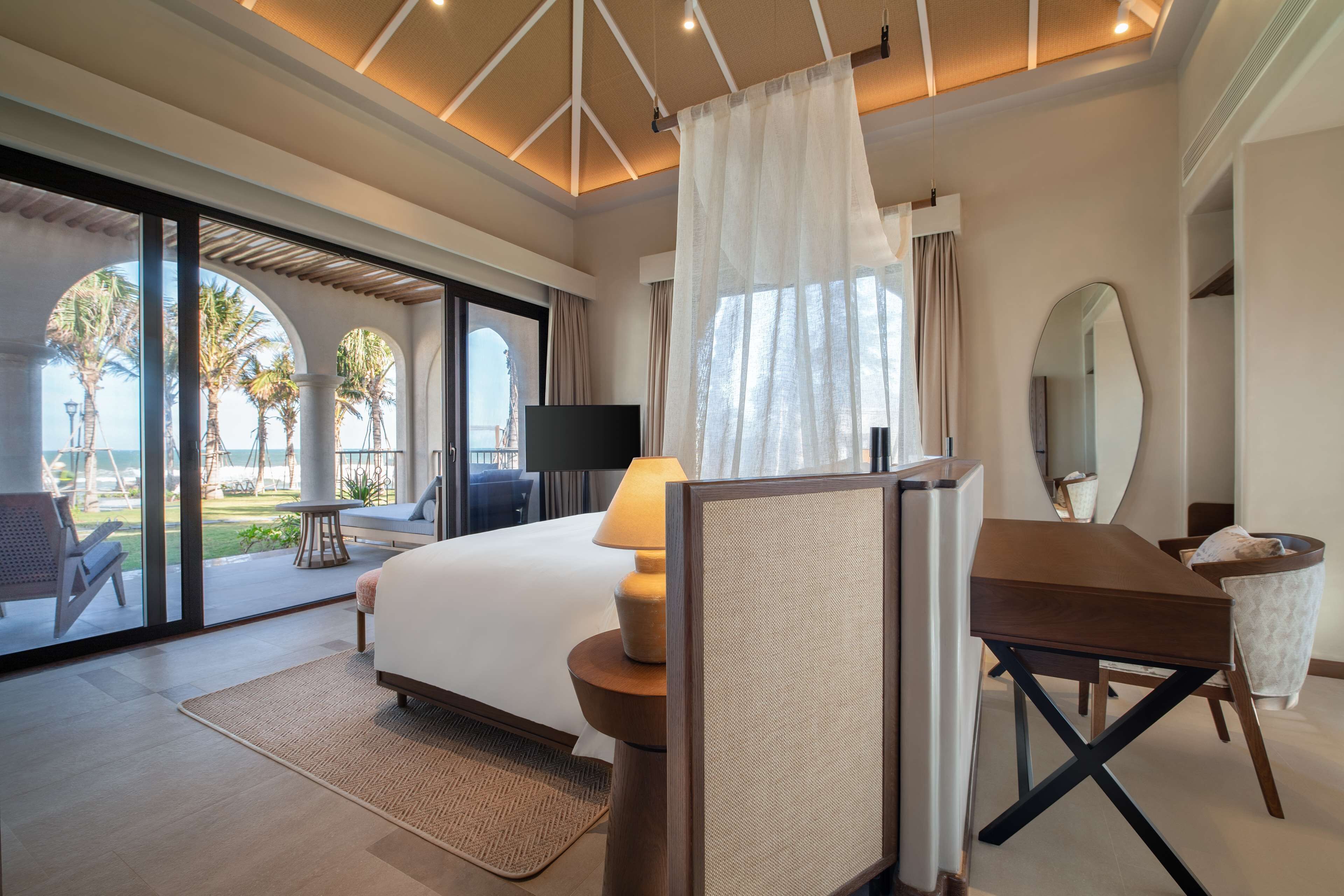 Une chambre d’hôtel équipée d’un lit, d’un bureau, d’une télévision et d’une grande fenêtre donnant sur des palmiers et une piscine.