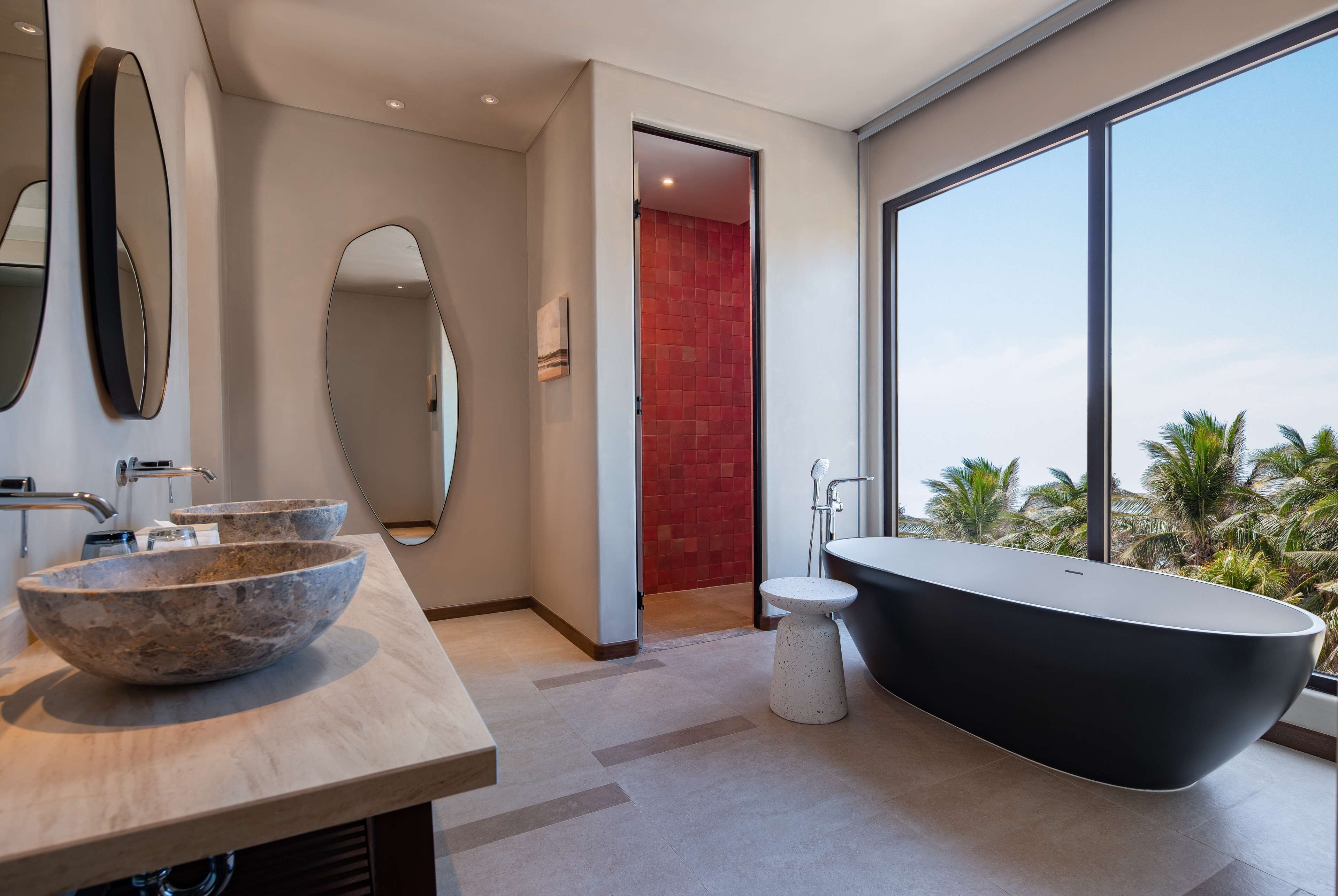 A modern bathroom with a freestanding bathtub, a large oval mirror, and a large window offering a view of palm trees.