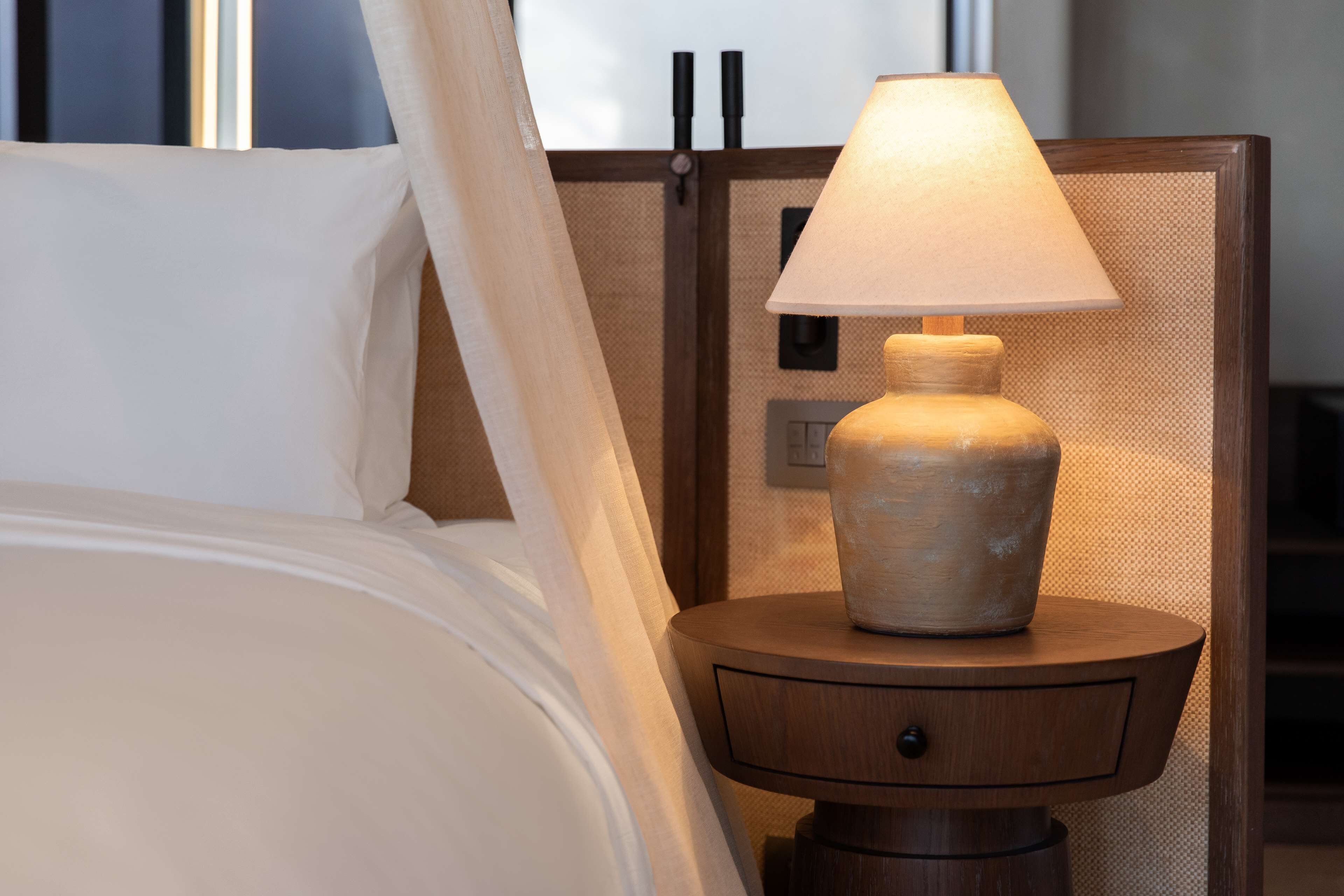 Een bed met witte lakens, een houten nachtkastje met een lamp, en een schakelaar aan de muur.