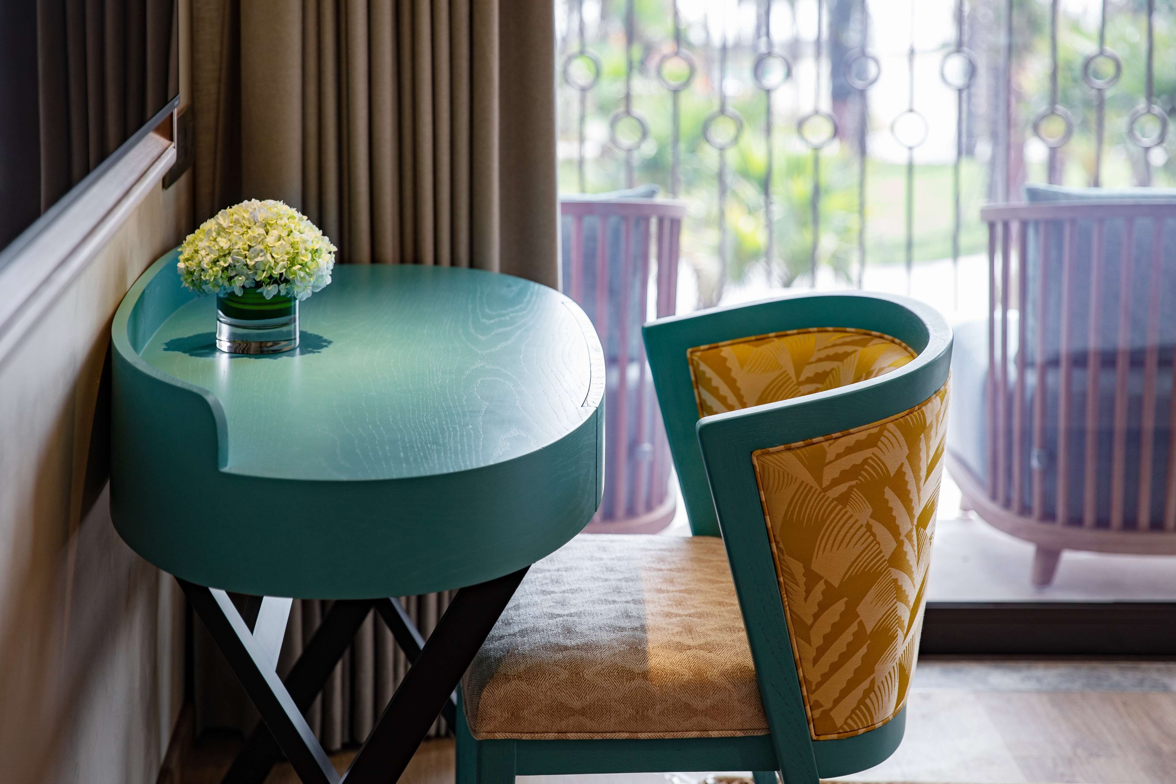 Una mesa redonda azul verdoso con un jarrón de flores blancas, una silla amarilla estampada y vistas a un balcón con barandillas de metal y follaje.