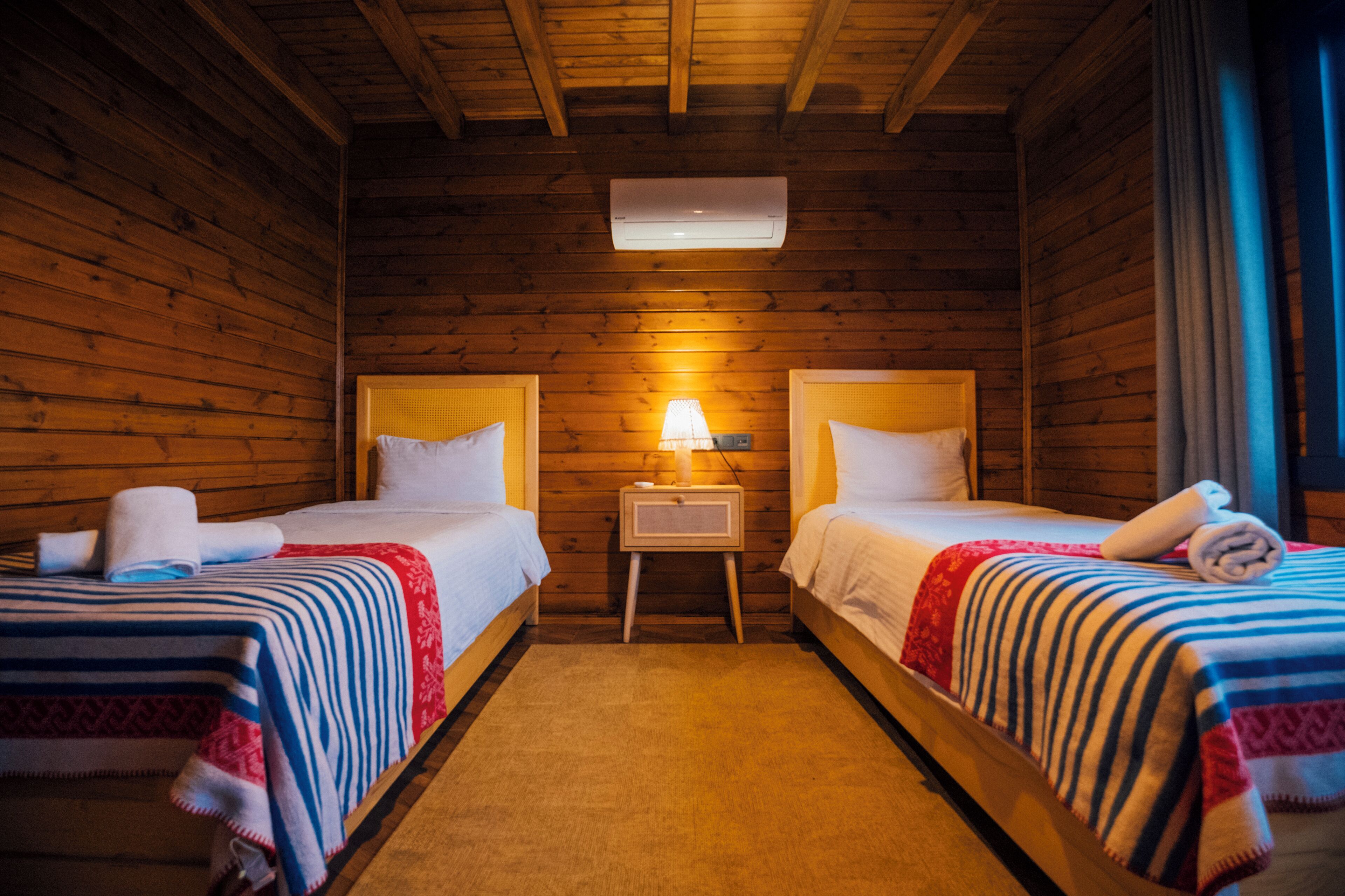 A cozy room with two beds, wooden walls, a small bedside table with a lamp, and an air conditioning unit.
