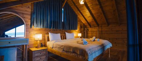 A wooden cabin bedroom with a bed, nightstand, lamp, and a view of the outdoors.