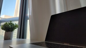 Um laptop sobre uma mesa perto de uma janela com vista para prédios e um vaso com planta.