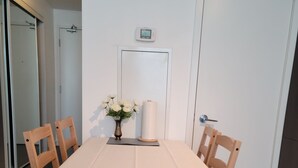 Uma área de refeições com uma mesa posta para seis pessoas, rodeada por cadeiras de madeira. Um vaso com flores brancas e um tapete enrolado estão sobre a mesa.