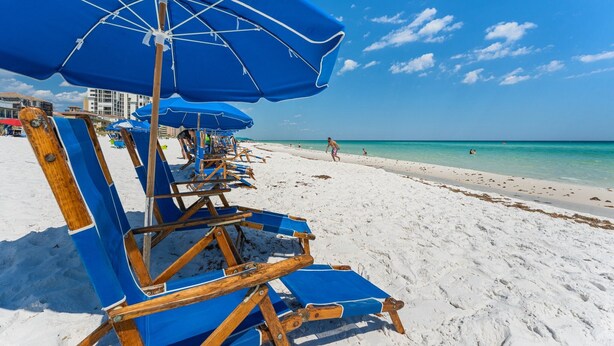 Uma praia com fileiras de cadeiras e guarda-sóis azuis, uma orla arenosa e um céu azul límpido.