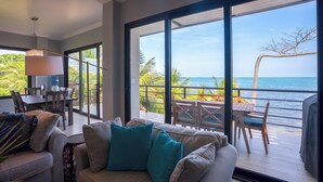 A modern living room with a large window overlooking a balcony with a view of the ocean. - Sunset Gecko Condos - Unit 1A (Placencia)