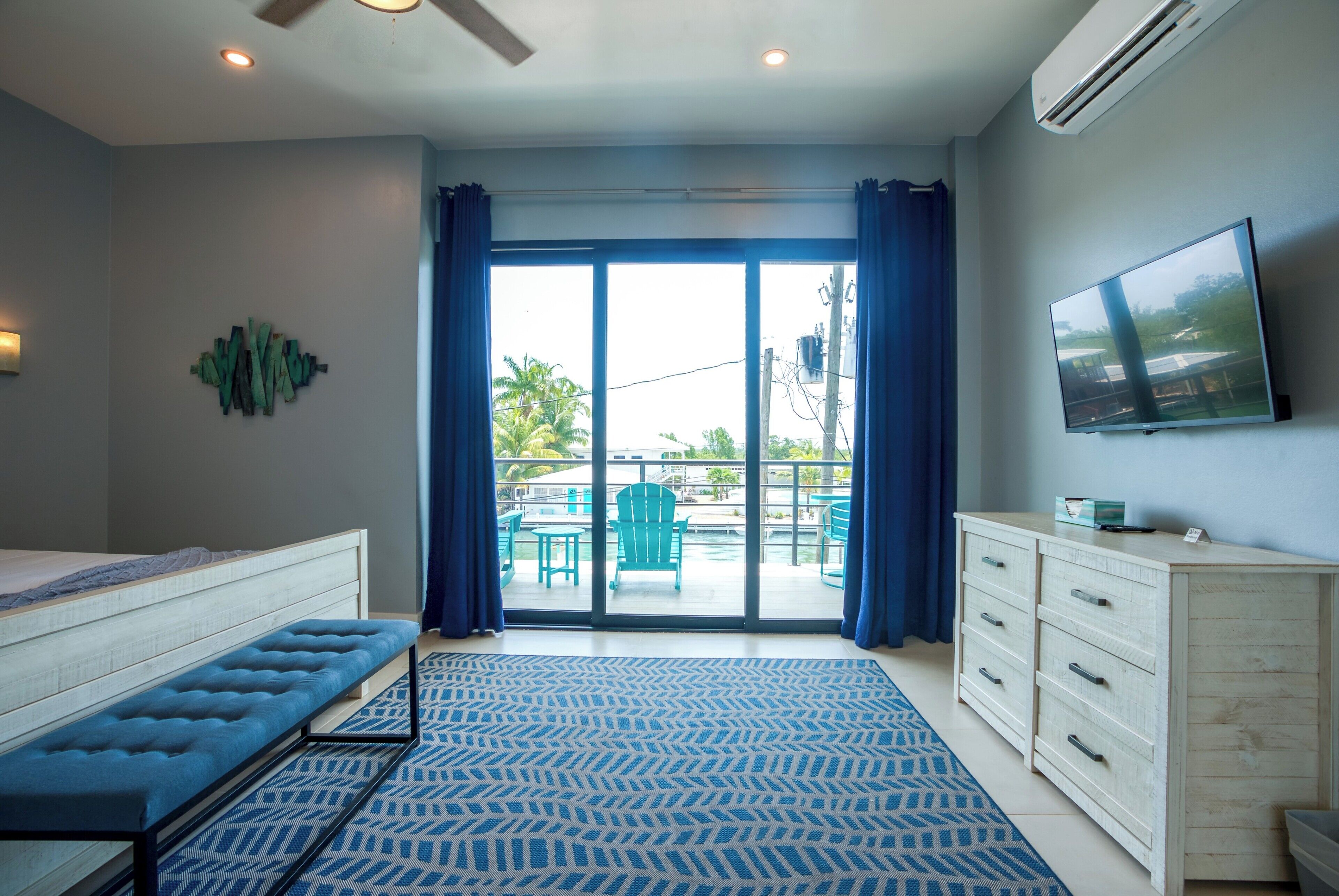 Een slaapkamer met een bed, een bankje, een dressoir, een televisie en uitzicht op een zwembad en terrasmeubels.