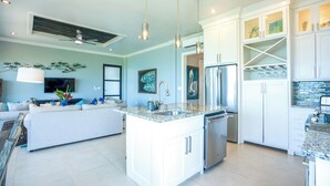 A modern kitchen with white cabinetry, a central island with a sink, and a dining area with a sofa and a TV. - Sunset Gecko Condos - Unit 1A (Placencia)